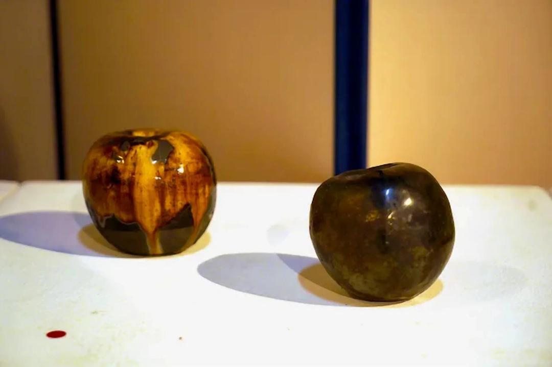 이동구, 〈사과〉, 도자, 9x9cm — 흰 선반 위에 사과 모양 도자가 둘 놓였고, 왼쪽은 호박빛 유약이 줄기처럼 흘러내리며, 오른쪽은 어두운 청동빛으로 기울어 있다