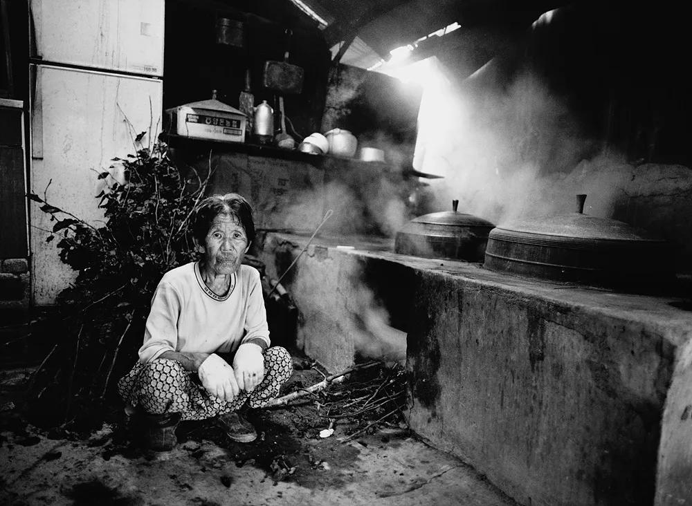 조문호, 〈2003정선〉, Pigment ink on FineArt Paper print, 70x50cm — 흑백 사진 속에서 왼쪽에 한 여성이 긴소매 상의와 무늬 바지 차림으로 쪼그려 앉아 카메라를 보고, 오른쪽 시멘트 부뚜막 위 솥뚜껑 둘에서 김이 오르며 위쪽 오른편으로 빛이 쏟아진다